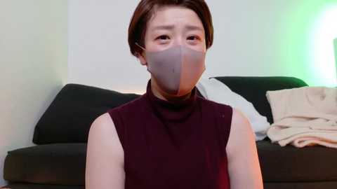 Media: Video of an Asian woman with short brown hair, wearing a maroon sleeveless top and a gray face mask, sitting on a black couch with white and beige pillows, against a light green wall.