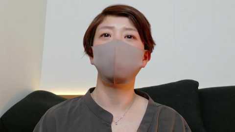 Media: Video of an East Asian woman with short brown hair, wearing a gray surgical mask, a gray t-shirt, and a necklace, sitting on a black couch against a light beige wall.