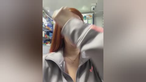 Media: Video of a woman with long red hair, wearing a gray hoodie, blurred and partially obscured, standing in a brightly lit pharmacy with shelves of products in the background.