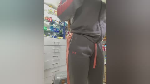 Video of a person's back in a gray Adidas track suit with red trim, hands in pockets, standing in a brightly lit convenience store.