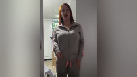 Video of a fair-skinned woman with long orange hair wearing a gray zip-up jumpsuit, standing in a narrow hallway with white walls.