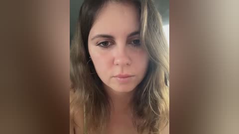 Media: Video of a young Caucasian woman with shoulder-length, wavy brown hair, fair skin, and neutral expression. She has a slender build and is likely in her early 20s. The background is out of focus, with a soft blur.