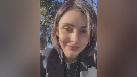 Media: Video of a young woman with shoulder-length dark hair, fair skin, and light makeup, smiling softly outdoors with sunlight filtering through trees in the background. She wears a dark blazer and a white shirt.