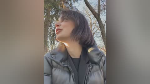 Media: Video of a young woman with short, dark hair, wearing a black leather jacket, standing outdoors in a forest. She has a serene expression and her head is tilted upward.