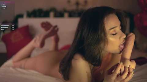 Video of a nude woman with long dark hair, lying on her stomach on a bed, giving a blowjob to a man's erect penis. Background shows a dimly lit bedroom with red pillows and a blurred view of a Christmas tree.