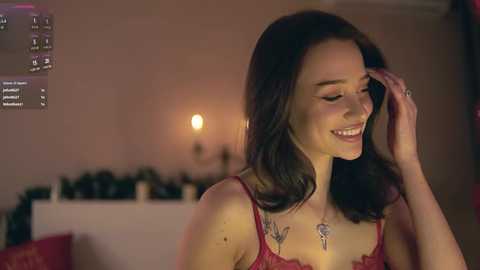 Video of a smiling young woman with shoulder-length dark hair, wearing a red lace bralette, standing in a softly lit room with a fireplace and holiday decorations.