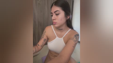 Media: A video of a young Latina woman with long dark hair, medium skin tone, and tattoos on her arms. She wears a white crop top and sits in a dimly lit room with a wooden door in the background.