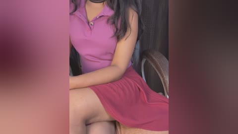 Media: Video of a woman with long, dark hair wearing a tight, sleeveless pink dress, sitting with her legs crossed on a wooden chair, revealing her bare thigh. Background is a dark wooden wall.