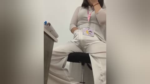 Media: Video of a woman in a white lab coat and pants sitting on a stool, wearing a lanyard with a badge, holding a clipboard, in a clinical setting.