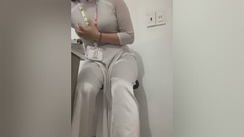 Media: A video of a woman in a white, long-sleeved, high-waisted dress, seated on a black chair. She holds a phone with a lanyard, against a plain white wall.