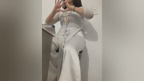 Media: A video of an Asian woman in a white lab coat and high-waisted pants, making a heart with her hands in front of a plain white wall.