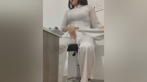 Media: A video shows a woman in white medical scrubs, seated on a stool, her legs spread, hand on her groin.