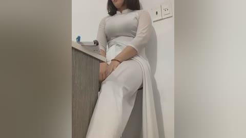 Media: Video of an Asian woman in a white, semi-sheer, long-sleeved dress, sitting on a counter in a minimalistic room with a light switch and a blue object on the countertop.