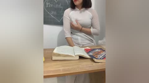 Media: Video of a woman in a white dress, standing at a wooden table with an open book and a chalkboard behind her.