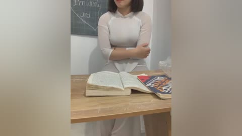 Media: Video of a woman in a white dress standing behind a wooden table in a classroom setting. The table has an open book and a colorful book with a cover featuring an orange background.