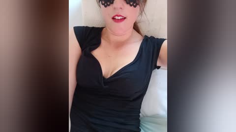 Media: Video of a fair-skinned woman with dark, curly hair and red lipstick, wearing a black, low-cut shirt and black lace mask, indoors with blurred background.