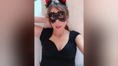 Media: Video of a fair-skinned woman with light brown hair, wearing a black lace mask with red devil horns, a low-cut black dress, and red lipstick.