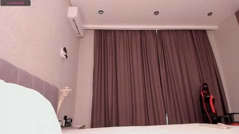 Video of a minimalist bedroom with a large, dark brown curtain covering the window, an air conditioner on the wall, a white chair, and a white bed with a light green pillow in the foreground.