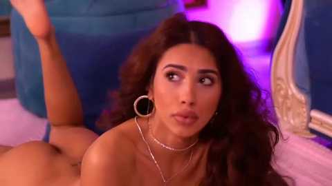 Media: Video of a topless, tan-skinned woman with long, dark brown hair, wearing large hoop earrings and a delicate necklace, lying on a blue cushioned surface under purple lighting, looking contemplative.