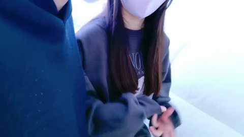 Media: Video of a masked woman in a gray sweater, with long brown hair, holding hands with a masked man in a dark blue sweater, standing in a brightly lit, modern room with white walls.