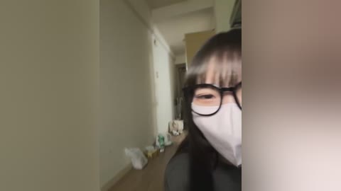 Media: A video of a woman with long, dark hair and glasses, wearing a white mask, partially visible in a dimly lit, narrow hallway with beige walls, a white door, and a cluttered countertop.