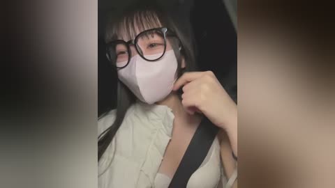 Media: Video of a young Asian woman with long black hair, wearing a white face mask, black-framed glasses, and a white blouse, seated in a car.