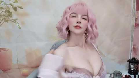 Media: Video of a pale-skinned woman with pink, wavy hair and large, bare breasts in a pastel-colored bra. She wears a blue cardigan, gold earrings, and stands against a soft-focus, floral background.