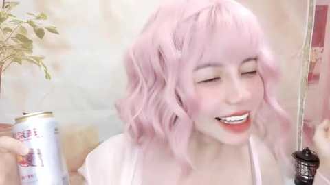 Media: Video of a smiling, fair-skinned woman with pastel pink wavy hair, wearing a white top, applying mascara in a bathroom with a plant and a can of hairspray.