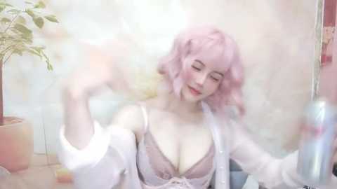 Media: A video of a pale-skinned woman with short, pastel pink hair, wearing a revealing white robe over a lavender lace bra, holding a can of spray. The background features a tiled wall and a potted plant.