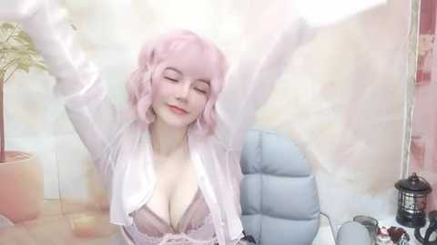 Media: Video of a smiling woman with pastel pink hair, wearing a white lace robe over a lacy pink bra, arms raised, sitting in a pastel room with plants and a blue chair.