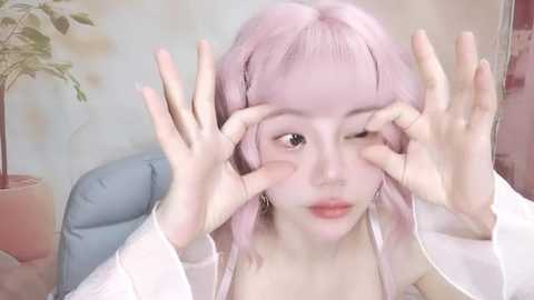 Media: Video of a fair-skinned, young Asian woman with pink hair, wearing a white robe, playfully covering her eyes with her hands. Background features a blue chair and a potted plant.