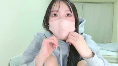 Media: A video of an East Asian woman with long black hair wearing a light pink face mask and a grey hoodie, sitting indoors with white walls and a bed visible in the background.