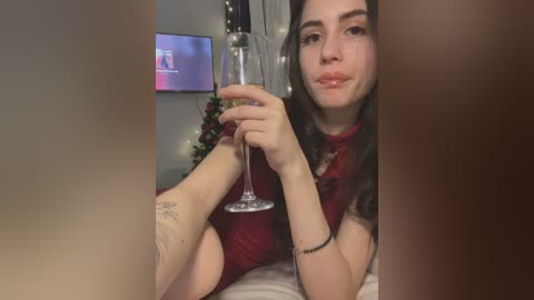 Media: Video of a young woman with fair skin, long dark hair, and light makeup, wearing a red dress, holding a wine glass, sitting on a bed, with a floral arrangement and TV in the background.