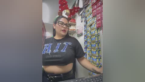 Media: Video of a Latina woman with medium skin tone, wearing glasses, black crop top with \"HIT\" in blue letters, and jeans, standing in a convenience store aisle.