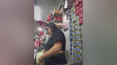 Media: Video of a Latina woman with long black hair and glasses, wearing a black shirt, shopping in a well-lit convenience store. She holds a bag of chips and looks at a display of chips and candy.