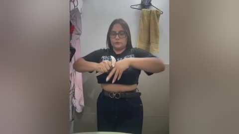 Media: Video of a young Latina woman with medium skin tone, dark hair, and glasses, wearing a black crop top and high-waisted dark pants, adjusting her bra in a bathroom with beige walls, clothes hanging on hooks, and a towel on a rack.