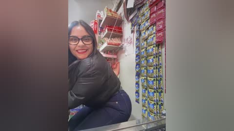 Media: A video of a Latina woman with long dark hair and glasses, wearing a black jacket and tight blue jeans, smiling while leaning over a counter in a convenience store.