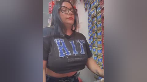 Media: Video of a woman with medium-sized breasts, wearing a black crop top with \"HIT\" in blue letters, and glasses, standing in a cluttered room filled with snack bags.