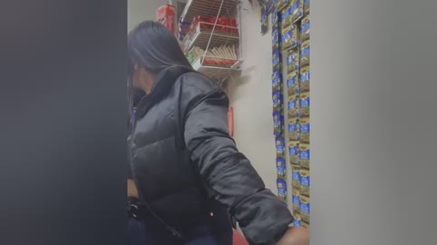 Media: Video of a person with long black hair, wearing a black puffer jacket, standing in a store with shelves of snacks, including chips and cookies, and a red display rack.