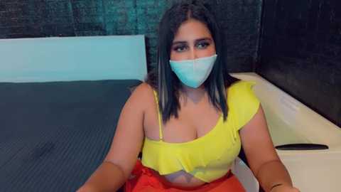 Media: Video of a plus-sized South Asian woman with dark hair, wearing a yellow cropped top, red skirt, and face mask, sitting on a massage table in a dimly lit room with dark walls.