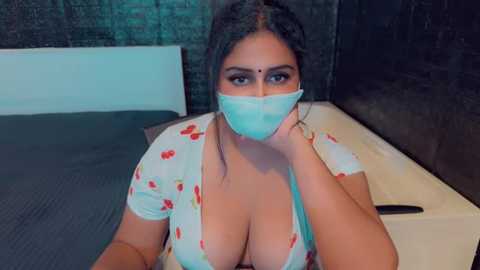 Media: Video of a South Asian woman with medium skin tone, wearing a blue surgical mask, floral-patterned white top, and red lipstick. She has a curvy physique, large breasts, and sits on a bed in a dimly lit room.