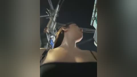 Media: A video of a woman in a dark room, facing away, with her head tilted back, wearing a black off-shoulder top. The background shows dimly lit industrial equipment, suggesting a laboratory or studio setting.