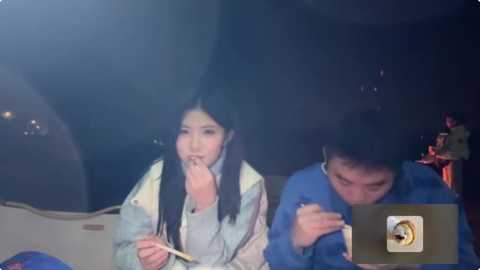 Media: Video of an Asian woman with long black hair, in a white jacket, and a man in a blue shirt, both holding chopsticks, eating noodles in a dimly lit restaurant.