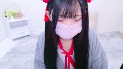 Media: Video of a young East Asian woman with long black hair, wearing a pink surgical mask, red ribbon, and a grey cardigan, kneeling on a grey carpeted floor in a minimalistic room with a white dresser and potted plants.