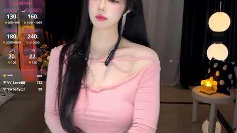 Media: Video of a fair-skinned woman with long black hair, wearing a pink off-shoulder top, seated in a dimly-lit room with a lit candle and colorful wall decor.