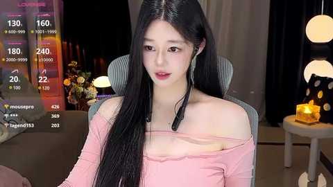 Media: Video of an East Asian woman with long black hair, wearing a pink off-shoulder top, sitting in a chair with a dimly lit, cozy room background.