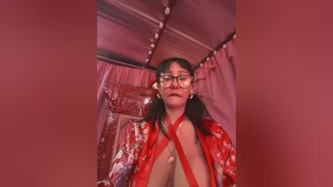 Media: A video of a young woman with long black hair and glasses, wearing a red kimono with floral patterns, sitting in a tent with pink curtains and white string lights.