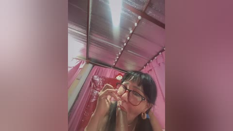 Media: Video of an Asian woman with long black hair, glasses, and large hoop earrings, smoking a cigarette in a dimly lit, pink-tinted room with reflective metal ceiling panels.