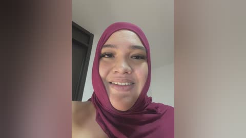 Media: Video of a smiling, dark-skinned woman with a medium build, wearing a magenta hijab and a matching top, in a dimly lit room with a white wall and a partially visible door.