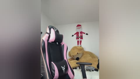 Video of a minimalist room with a black-and-pink gaming chair, a large teddy bear, and a cartoonish Santa Claus decoration on a white wall.